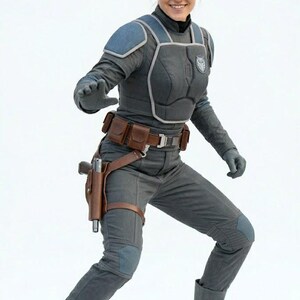 May include: A person in a gray and blue armored costume with a utility belt and holster. The costume includes a chest plate, shoulder pads, gloves, and boots. The person is smiling and posing against a white background.