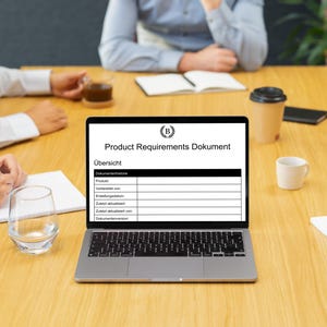 May include: A laptop displaying a document titled "Product Requirements Document" is open on a wooden table. The document has a black and white table with several blank rows. Several people are seated around the table, with coffee cups and notepads visible.