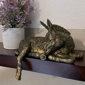 May include: A bronze-colored donkey figurine in a resting pose. The sculpture has detailed features and is positioned on a dark brown shelf. A potted lavender plant is in the background.
