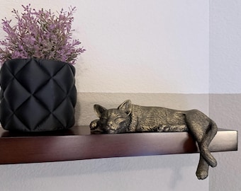 May include: A bronze-colored cat figurine in a sleeping position with its tail and hind legs hanging over the edge of a dark brown shelf. Next to the cat is a black, quilted-texture cylindrical planter containing a small, light purple plant.