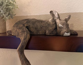 May include: A brown rabbit figurine with textured fur, depicted in a resting position with eyes closed. The rabbit features one long hind leg extending downward over the edge of a surface, while the rest of the body lies flat along the top edge.