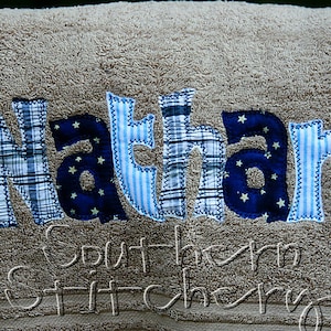 May include: A beige towel with the name "Nathan" spelled out in blue and white fabric letters. The letters are sewn together with a zig-zag stitch. The text "Southern Stitchery" is embroidered in white on the bottom of the towel.