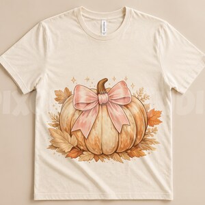 Peut inclure: Un t-shirt à manches courtes de couleur crème arborant un motif central représentant une citrouille orange clair ornée d'un grand nœud rose pâle. La citrouille est entourée de feuilles d'automne dans des tons orange et marron, avec des étoiles.