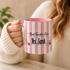 May include: A ceramic mug with a pink and white vertical striped pattern. The text "Best Teacher Ever" and "Mrs. Sarah" is printed in black, accompanied by small illustrations of two yellow pencils and two red apples on the front surface.