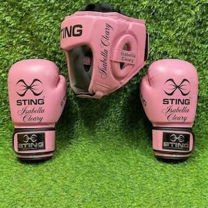 Op de afbeelding: Een set roze boksuitrusting bestaande uit twee bokshandschoenen en een hoofdbeschermer. Elk item is voorzien van het zwarte Sting-logo, de naam Isabella Cleary in cursief schrift en een klein rechthoekig label met de naam Isabella Cleary.