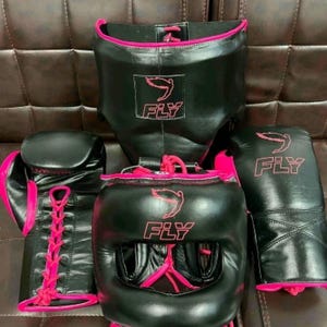 Può includere: Un set da boxe nero con dettagli rosa, tra cui caschetto, guanti e paradenti. Ogni pezzo presenta il logo "FLY" in rosa. I guanti hanno lacci rosa.