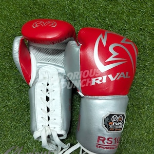 May include: Pair of red and silver boxing gloves with white laces. The gloves have the word "RIVAL" in silver on the red portion. The gloves are on a green surface. The text "RS10" and "SPARRING GLOVES" is visible.