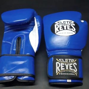 Pode incluir: Um par de luvas de boxe azuis com detalhes brancos e acabamentos pretos. As luvas apresentam o logotipo "CLETO REYES" em branco e dourado. As luvas são feitas de couro e são projetadas para uso profissional.
