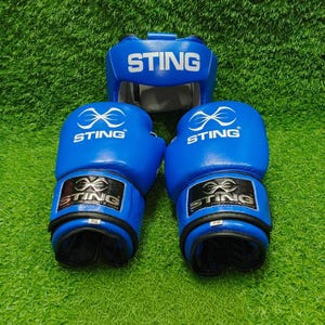 Op de afbeelding: Een blauwe boksset bestaande uit een hoofdbeschermer en twee bokshandschoenen van 14 ounce. Elk item is voorzien van het witte STING-logo en de tekst. De handschoenen hebben zwarte polssluitingen en de hoofdbeschermer heeft een STING-label.