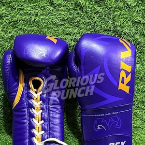 Könnte beinhalten: Paar lila Boxhandschuhe mit goldenen Akzenten und weißen Schnürsenkeln. Die Handschuhe tragen die Aufschrift "Glorious Punch" und "Rival". Der Hintergrund ist grün.