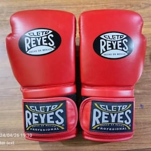 Könnte beinhalten: Paar rote Boxhandschuhe mit dem Markennamen "CLETD REYES" in weißer Schrift auf einem schwarzen Oval. Die Handschuhe tragen auch die Aufschrift "HECHO EN MEXICO" und "PROFESIONAL".