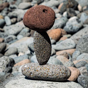 Könnte beinhalten: Ein ausgewogener Stapel von Flusssteinen. Die Basis ist ein großer, grauer Stein, der einen gesprenkelten, ovalen Stein trägt. Ein strukturierter, länglicher Stein ist zentriert, gekrönt von einem rötlich-braunen, abgerundeten Stein.