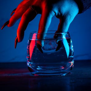 Könnte beinhalten: Ein Glas Wasser, in das eine Hand greift, beleuchtet von rotem und blauem Licht. Die Hand hat lange, schwarze Fingernägel und einen Ring. Der Hintergrund ist dunkelblau und erzeugt eine dramatische und geheimnisvolle Atmosphäre.
