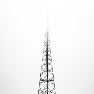 Könnte beinhalten: Ein hoher, schwarzer Funkmast vor einem weißen, nebligen Himmel. Die filigrane Gitterstruktur des Turms verjüngt sich nach oben und verschwindet im Nebel. Das Bild ist ein Spiel der Kontraste, mit dem dunklen Turm vor dem hellen Hintergrund.