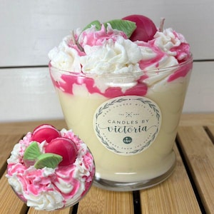 May include: A decorative candle in a clear glass, resembling a dessert. It features a creamy yellow wax base, topped with white and pink swirls, and decorative pink and green accents. The candle has a label that reads "Candles by Victoria."