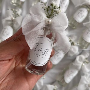 May include: Clear glass spray bottles with white labels, tied with white bows and adorned with small white flowers and pearls. The label reads "T & B" and "19.05.2024". The bottles are likely wedding favors or gifts.