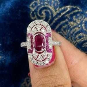 May include: An elongated oval ring featuring a central oval-cut deep red ruby surrounded by a geometric silver-toned setting. The frame is accented with smaller square-cut rubies and numerous tiny clear white stones set in a symmetrical pattern.