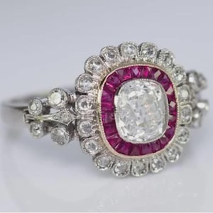 May include: A vintage-style ring featuring a large, square-cut, clear gemstone at the center, surrounded by a halo of smaller, round, clear stones and a border of red gemstones. The band is silver-toned with delicate detailing.