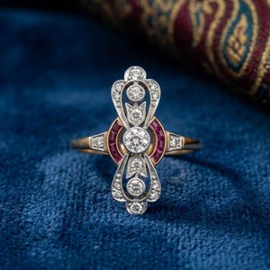 Può includere: Un anello in oro con un elemento centrale verticale allungato incastonato con molteplici diamanti rotondi taglio brillante e due rubini curvi taglio baguette. La struttura metallica presenta filigrana argentata e bordi milgrain.