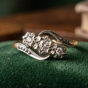 May include: A gold ring featuring three round diamonds set in a horizontal row. Each diamond is encased in a star-shaped metal setting. The band displays a curved, bypass-style design with intricate milgrain detailing along the edges.