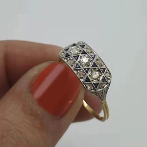 Puede incluir: Un anillo de estilo vintage con una cara rectangular. El anillo tiene una banda dorada y una parte superior plateada adornada con diamantes y piedras preciosas azul oscuro en un patrón geométrico. El anillo lo sostiene una persona con esmalte de uñas rojo.
