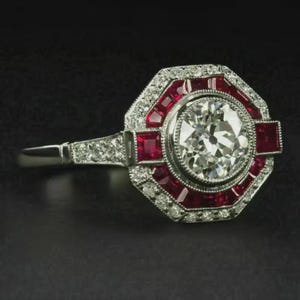 Może przedstawiać: Platynowy pierścionek z dużym, okrągłym, przezroczystym diamentem w środku. Pierścionek posiada otoczkę z małych diamentów i czerwonych kamieni szlachetnych, z dodatkowymi diamentami na obrączce. Pierścionek ma wzór Art Deco.