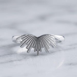 May include: A silver-toned ring featuring a central decorative element shaped like a sunburst or fan with multiple radiating rays. The metal has a smooth, polished finish, and the design is symmetrical, extending upward from the band in a V-shape.