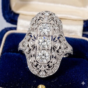 May include: A silver-toned ring featuring three prominent round-cut stones arranged vertically in a central rectangular setting, surrounded by an intricate filigree metalwork frame encrusted with numerous small, sparkling round-cut stones.
