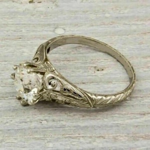 May include: An antique-style engagement ring with a large, clear gemstone. The ring has detailed filigree work on the band and around the gemstone setting. The metal appears to be silver or platinum.
