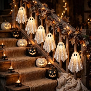 Peut inclure: Une guirlande de six lumières en forme de fantômes blancs à paillettes suspendue à une rampe d'escalier en bois décorée de feuilles d'automne et de pommes de pin. Des citrouilles illuminées et des bougies sont posées sur les marches.
