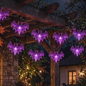 Può includere: Una catena di luci LED a forma di pipistrello viola appesa a un pergolato in legno. I pipistrelli in plastica traslucida sono illuminati dall'interno, emettendo una luce vivace. La catena luminosa presenta diverse figure di pipistrello.