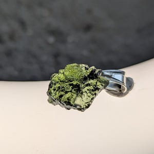 May include: A green and black moldavite pendant with a silver-coloured bail. The natural stone has a rough, textured surface. The pendant is displayed on a white surface.