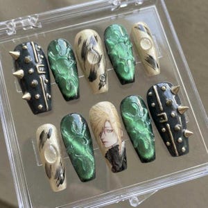 Op de afbeelding: Een set van tien kunstnagels in een doorzichtige plastic doos. De nagels hebben verschillende ontwerpen, waaronder zwarte en gouden studs, groene wervelpatronen en anime-personagekunst. De nagels hebben een doodskistvorm.