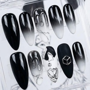 May include: A set of ten artificial nails in a clear plastic case. The nails feature various designs, including solid black, black to white ombre, and artistic drip and graphic designs. One nail has a silver cube charm.