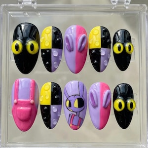 May include: A set of ten artificial nails in a clear display case. The nails feature various designs, including black with yellow eyes, split color patterns of yellow and black, and purple with pink accents. One nail has a cartoon bunny design.