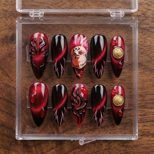 May include: A set of ten press-on nails in a clear plastic case. The nails feature a variety of designs, including red and black swirls, a face design, and gold accents. The nails are almond-shaped and have a glossy finish.