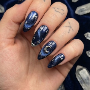 May include: Dark blue, almond-shaped nail design with silver accents. The nails feature a starry night effect with silver star and crescent moon embellishments. The design is set against a dark blue velvet background.