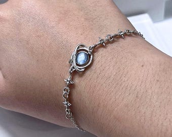 May include: A silver bracelet featuring a central blue gemstone encircled by a decorative setting. The bracelet's chain is adorned with star-shaped accents, creating a celestial theme. The bracelet is designed to be worn on the wrist.