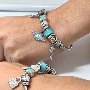 May include: Two silver charm bracelets with various charms, including a blue-green bead, a key, a four-leaf clover, and an evil eye charm. The bracelets are on a person's wrist.
