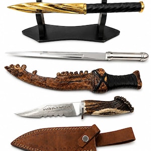 May include: A collection of items including a gold and black spiral-patterned wand on a black stand, a silver letter opener, a curved brown textured sheath, a serrated knife with an antler handle and etched text, and a brown leather sheath.