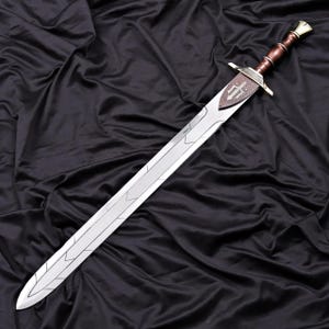 Pode incluir: Uma espada longa e prateada com uma guarda decorativa e um cabo envolto em couro castanho. A l&acirc;mina apresenta desenhos gravados e uma guarda em forma de escudo com um bras&atilde;o. A espada &eacute; exibida sobre um fundo preto.