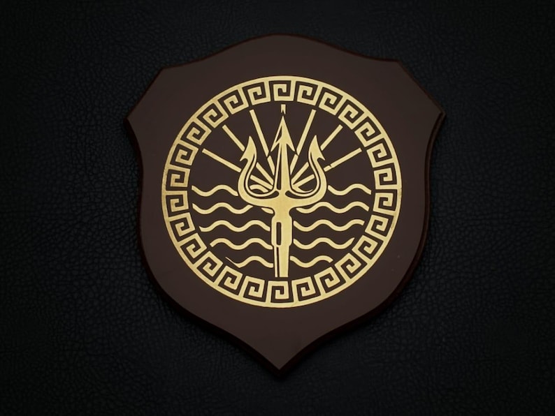 Pode incluir: Placa de madeira castanha com um emblema dourado com um tridente, ondas e um desenho de raios de sol dentro de uma borda de chave grega. A placa tem formato de escudo e est&aacute; sobre um fundo escuro e texturizado.