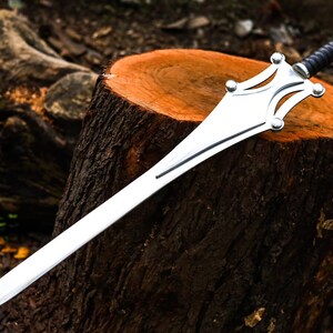 Op de afbeelding: Een zilveren zwaard met een zwart lederen handvat rust op een boomstronk. Het lemmet is lang en recht, met een decoratieve pareerstang met afgeronde accenten. Het zwaard is een fantasy-item.