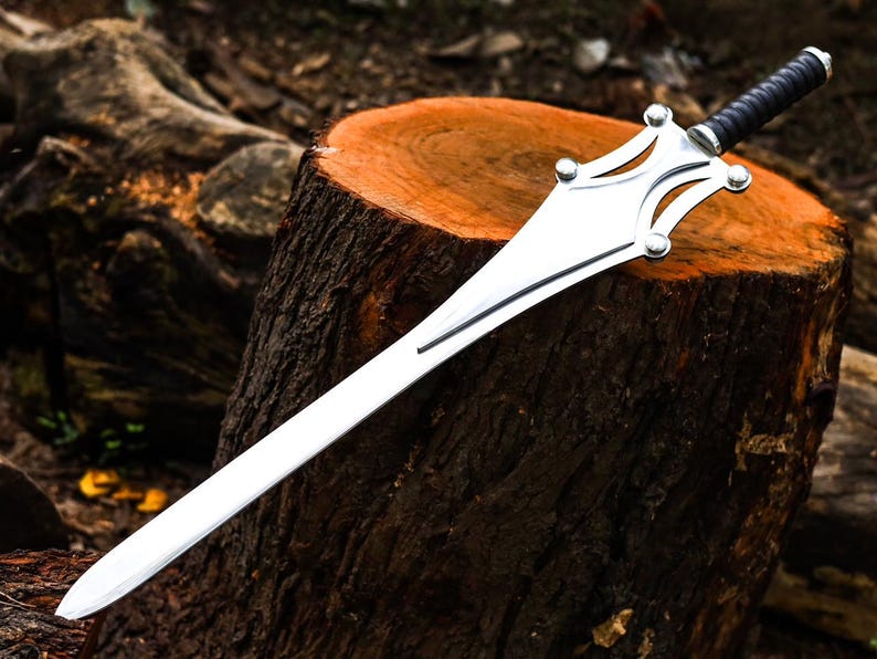 Op de afbeelding: Een zilveren zwaard met een zwart lederen handvat rust op een boomstronk. Het lemmet is lang en recht, met een decoratieve pareerstang met afgeronde accenten. Het zwaard is een fantasy-item.