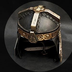 Peut inclure: Casque de style médiéval avec une base en cuir noir et des ornements en métal doré. Le casque présente un motif en forme de croix sur le dessus et une bande décorative autour de la base, avec une jugulaire.