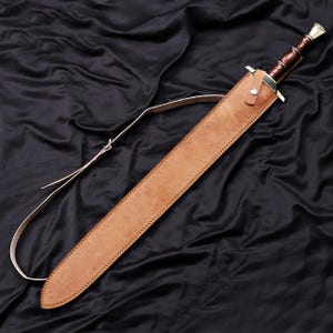 Pode incluir: Uma espada com bainha e correia de couro castanho, sobre um fundo preto. A espada tem uma l&acirc;mina prateada e um cabo castanho. A bainha &eacute; bege com costuras.