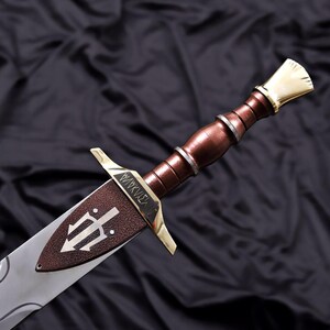 Pode incluir: Espada detalhada com punho castanho e l&acirc;mina prateada. A guarda apresenta detalhes dourados e uma guarda castanha com um s&iacute;mbolo de tridente. A l&acirc;mina tem um design curvo. A espada est&aacute; sobre um fundo escuro.