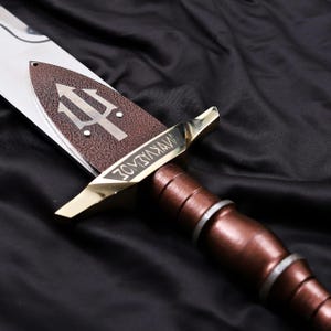 Pode incluir: Uma espada decorativa com cabo castanho e l&acirc;mina bronzeada em close-up. A l&acirc;mina apresenta um s&iacute;mbolo de tridente e a palavra "WYKVIADZ" gravada na guarda. A espada est&aacute; sobre um fundo preto.