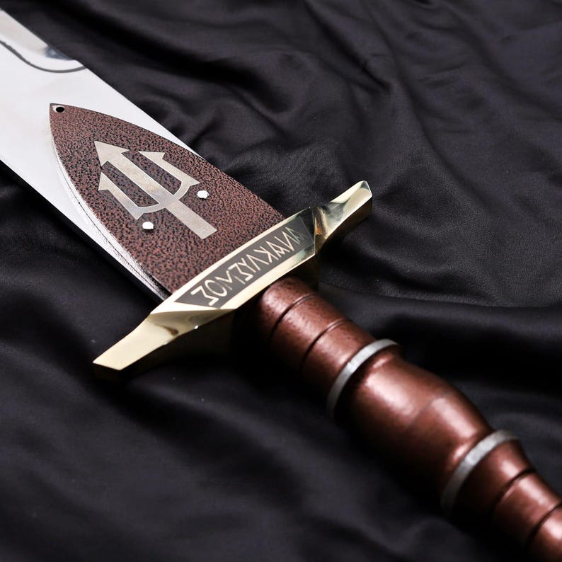 Pode incluir: Uma espada decorativa com cabo castanho e l&acirc;mina bronzeada em close-up. A l&acirc;mina apresenta um s&iacute;mbolo de tridente e a palavra "WYKVIADZ" gravada na guarda. A espada est&aacute; sobre um fundo preto.