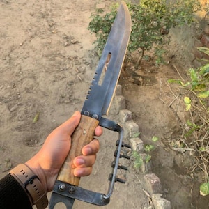 May include: A large, bladed knife with a wooden handle and a metal guard. The blade is dark gray, and the handle is light brown. The guard has a loop and a series of bolts. The knife is held in a hand.